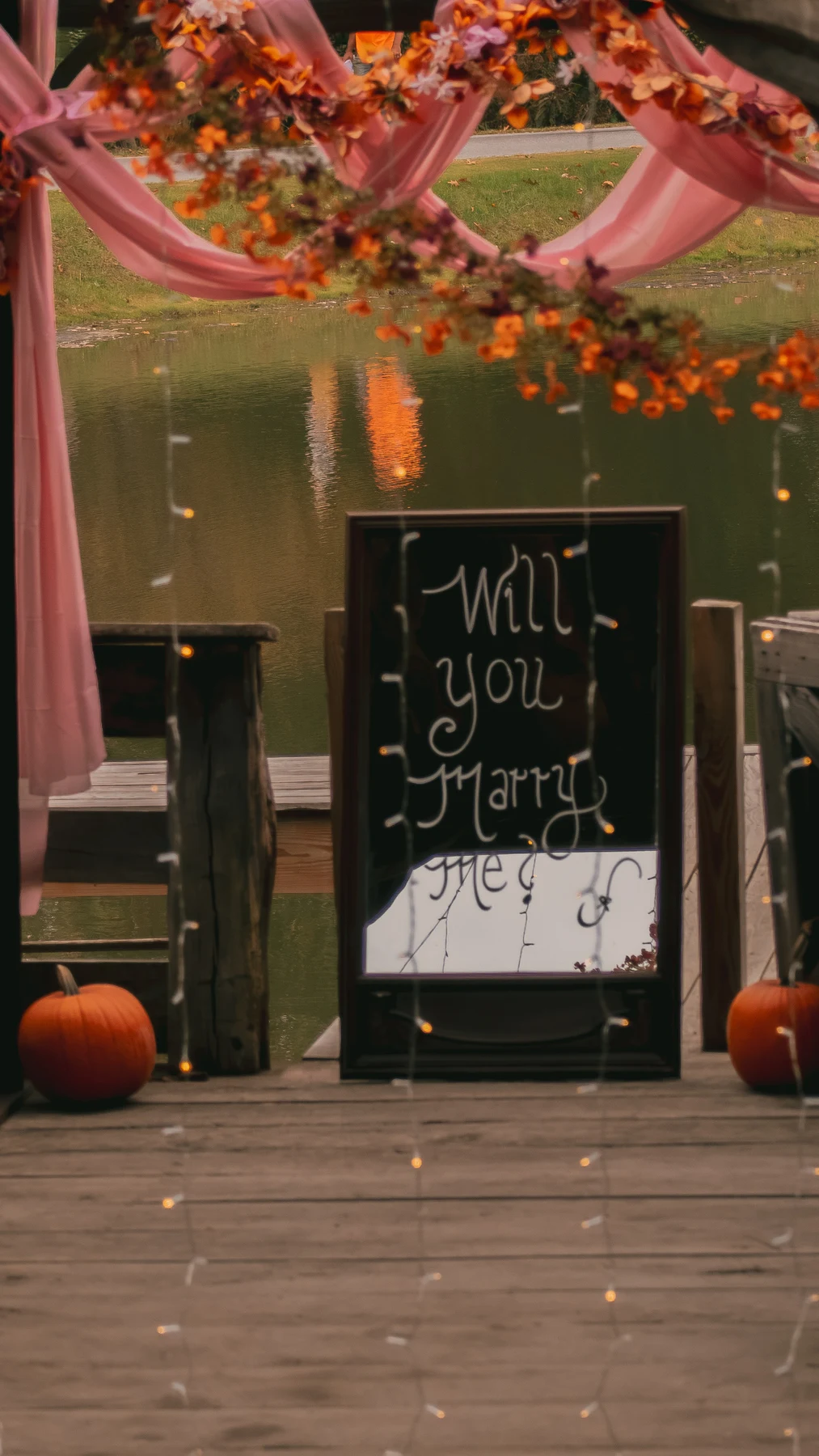 Will You Marry Me mirror framed by autumn flowers and hanging lights beside the pond