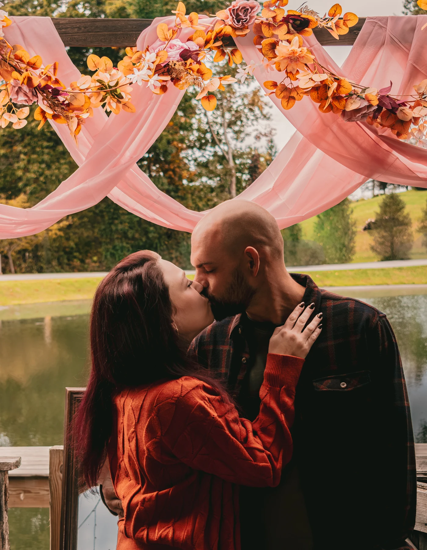 Couple kissing beneath pink drapery and autumn flowers beside a pond