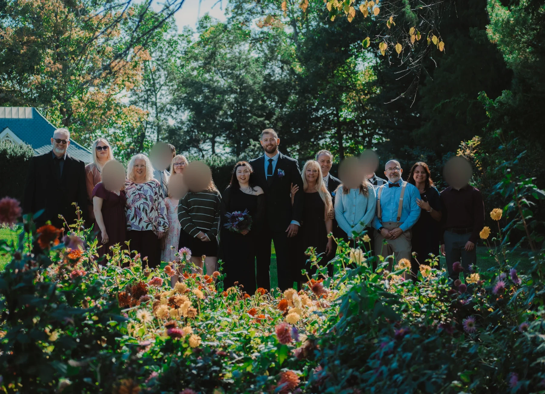 Emi and Mike with their wedding guests behind colorful garden flowers, with younger guests' faces blurred for privacy