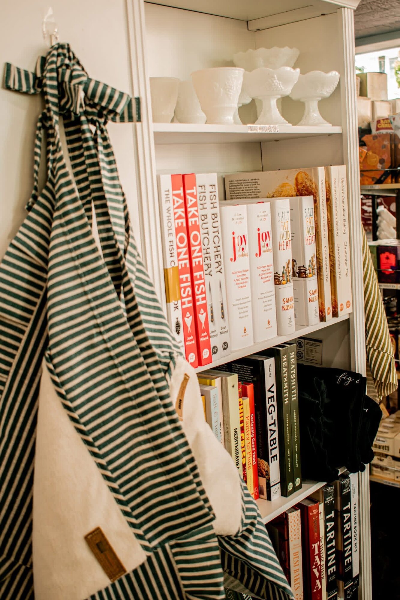Striped market aprons hanging beside a shelf of cookbooks and milk glass