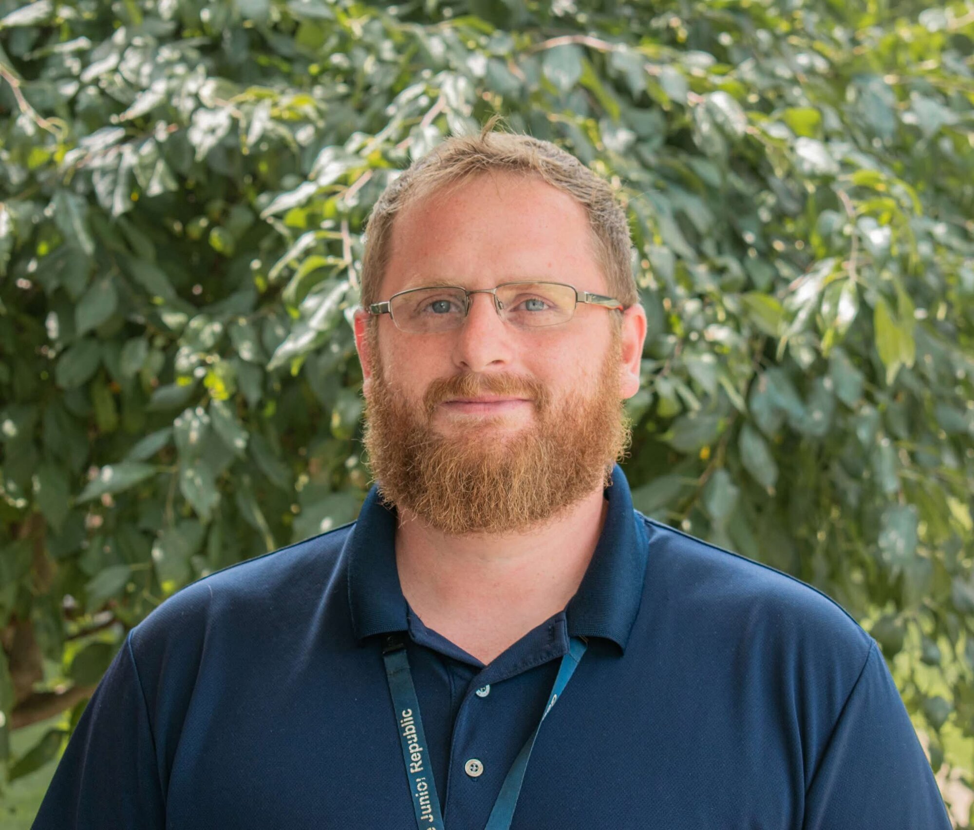 Outdoor professional headshot of a bearded man in a navy polo against greenery