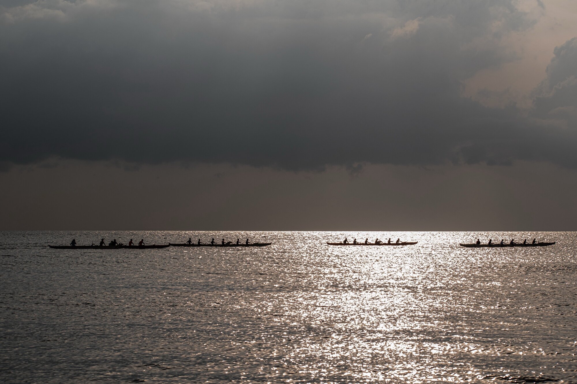 Outrigger canoes crossing silver water beneath a storm sky