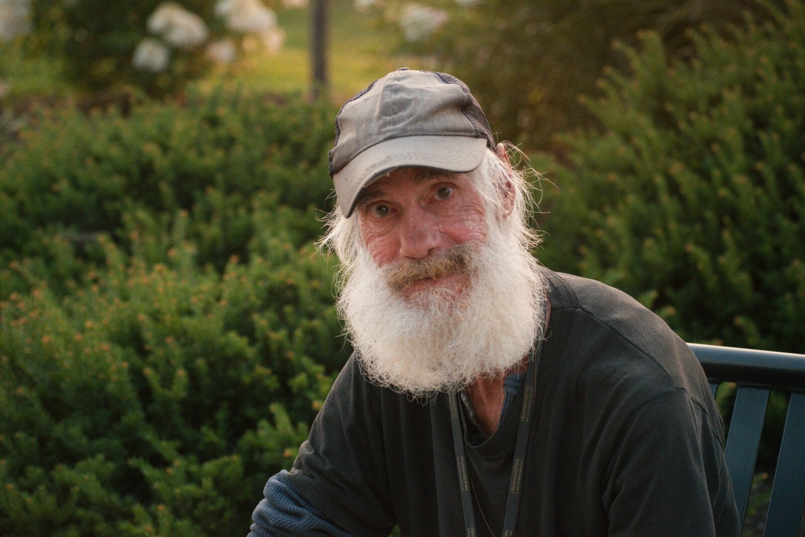 Golden-hour environmental portrait of a bearded man in a trucker cap looking at the camera among evening greenery
