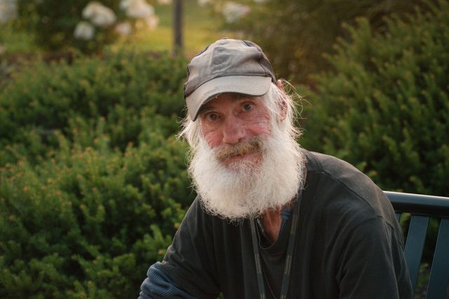 Golden-hour environmental portrait of a bearded man in a trucker cap looking at the camera among evening greenery