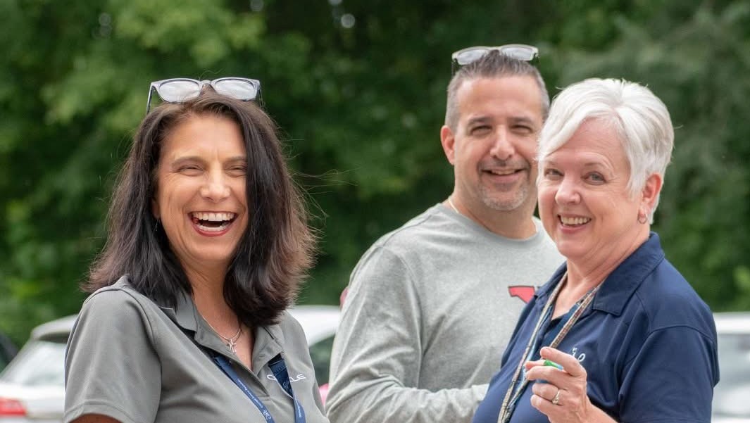 Three adult staff members laughing together outdoors at a community event
