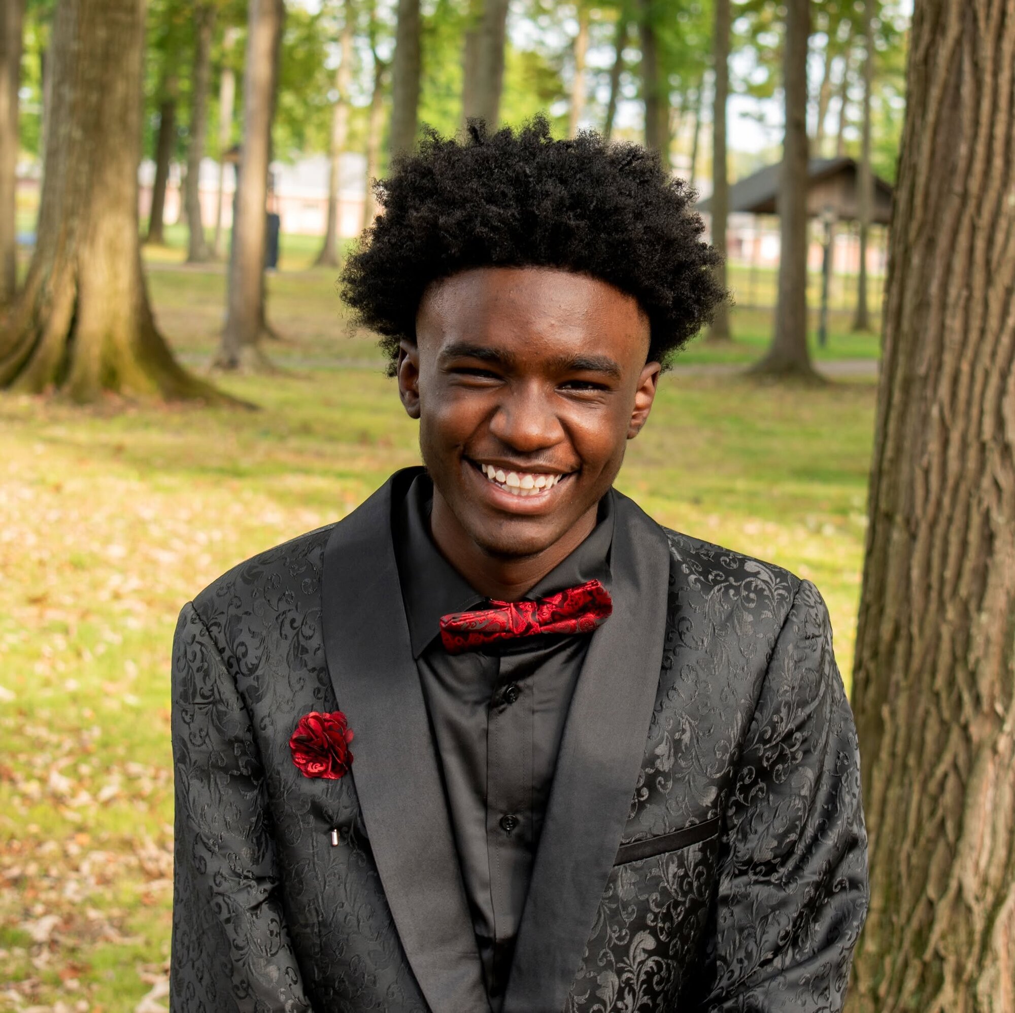 Smiling senior portrait of an adult in a patterned black suit jacket in a wooded park