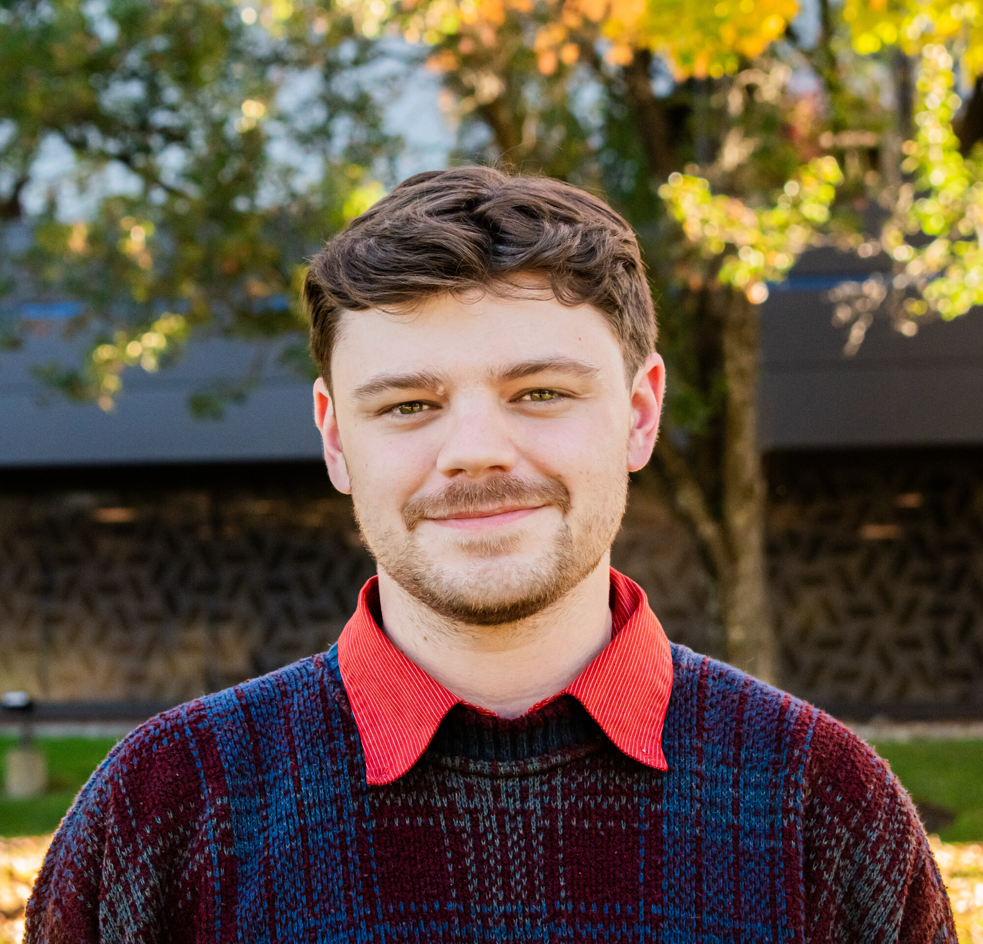 Outdoor headshot in a patterned sweater and red collared shirt, with autumn foliage behind