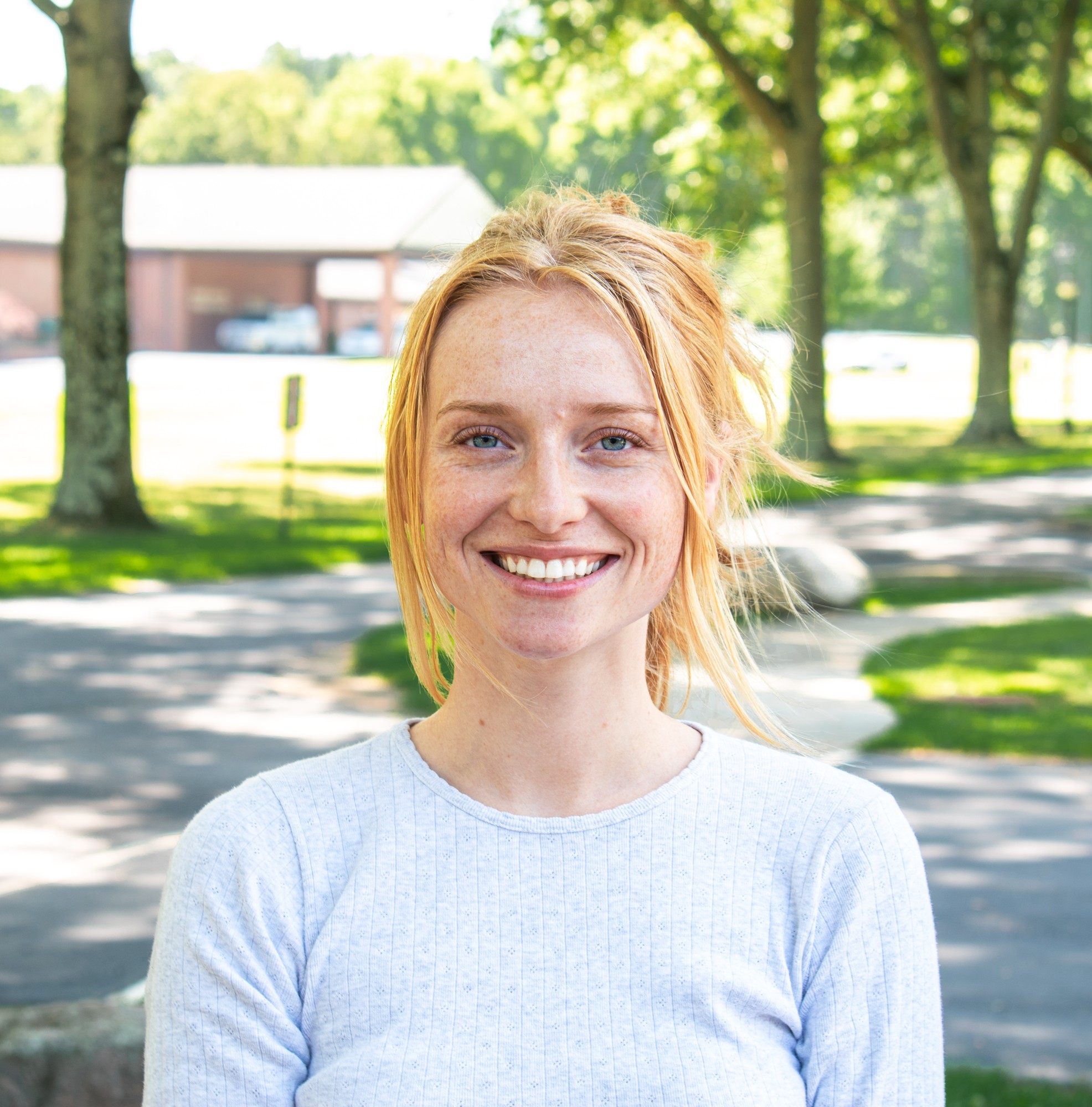 Outdoor portrait of a smiling red-haired woman in a light gray top beneath sunlit trees