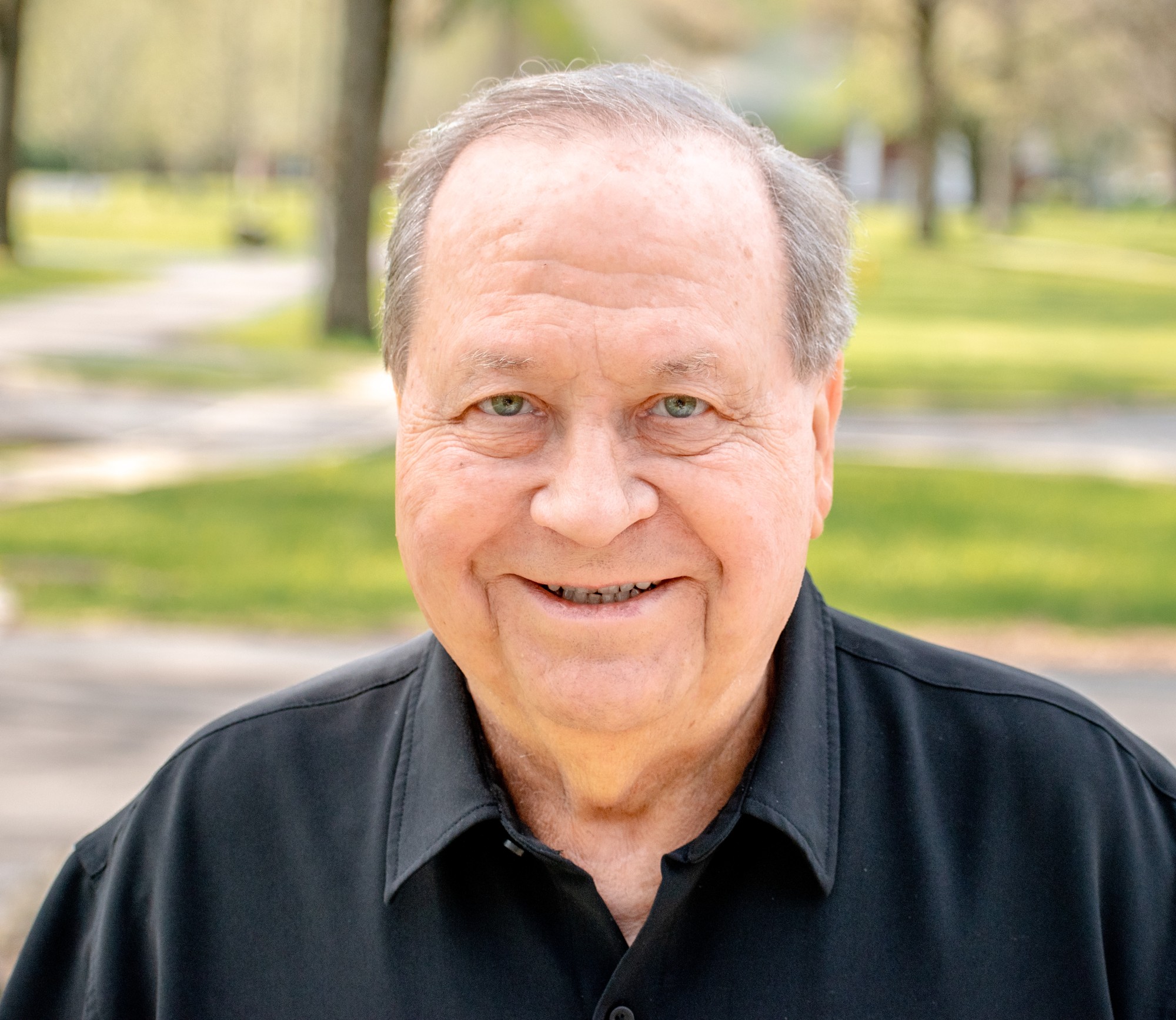 Outdoor headshot of a smiling man in a black collared shirt against softly blurred trees