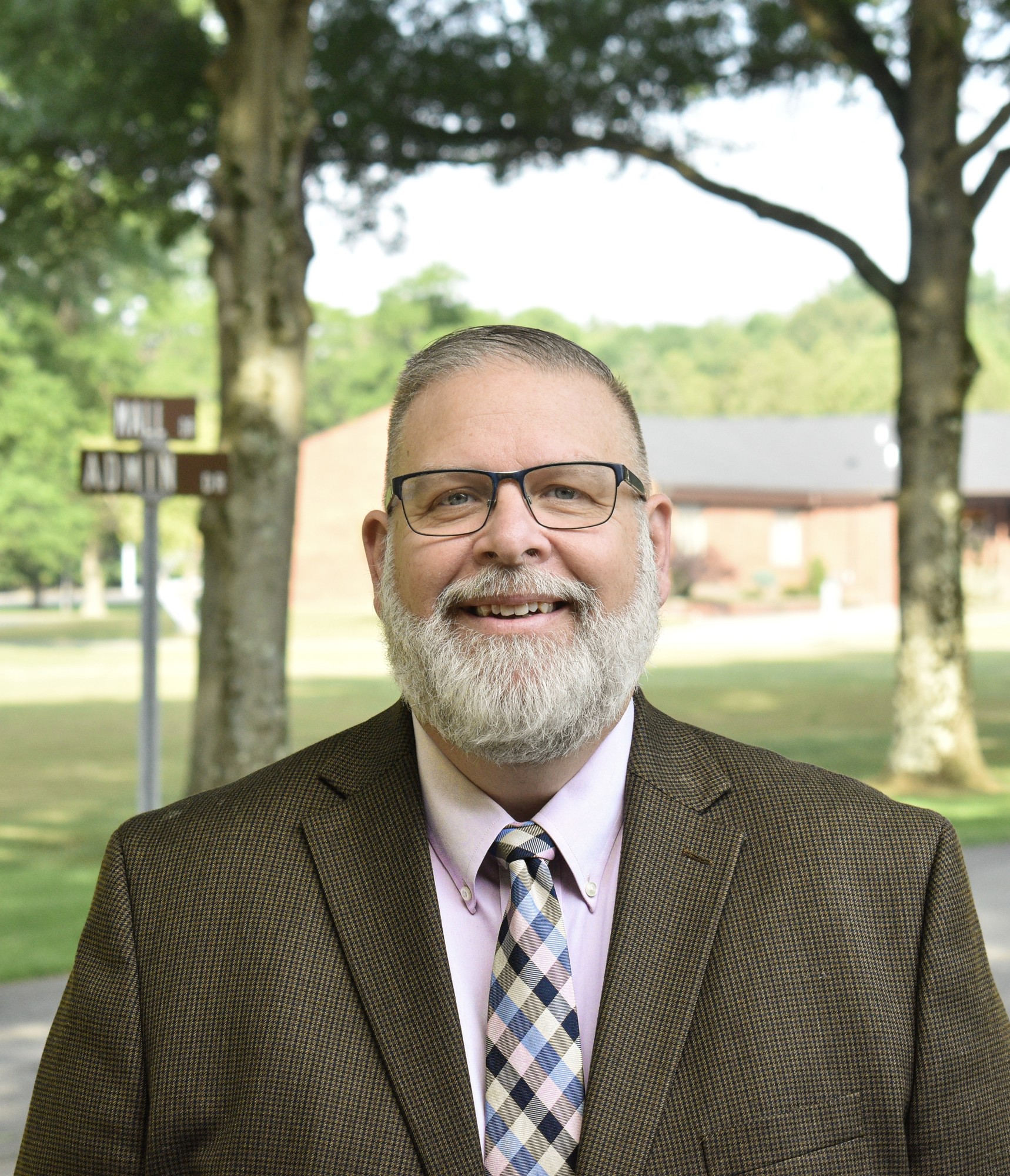 Outdoor professional portrait of a smiling man in glasses, a brown jacket, and a patterned tie beneath leafy trees