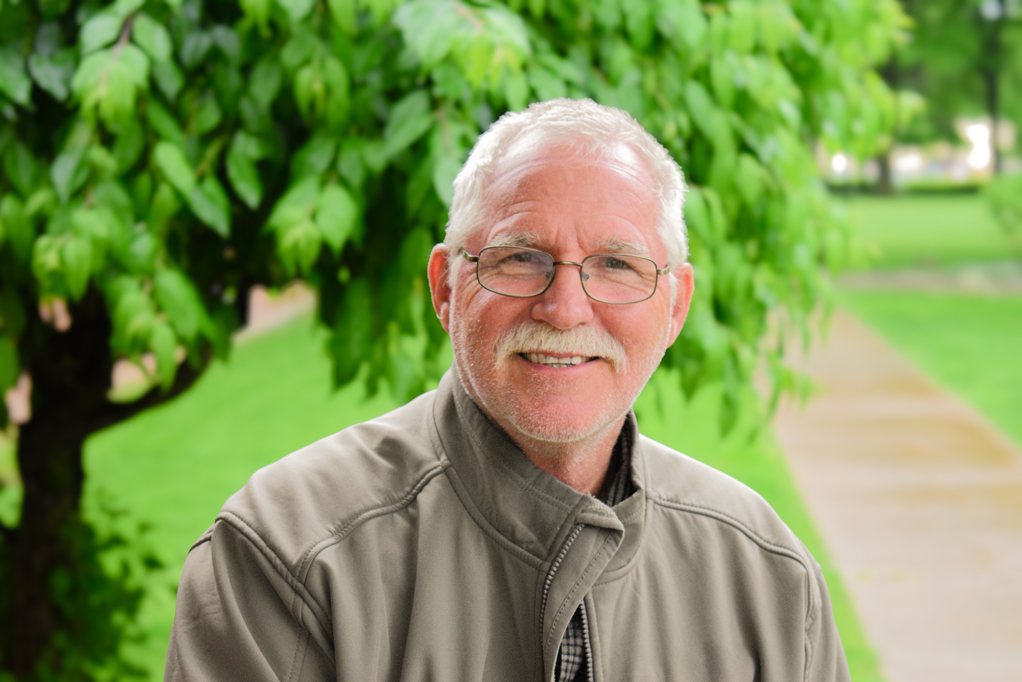 Outdoor portrait of a smiling white-haired man in glasses and a gray jacket beneath green leaves