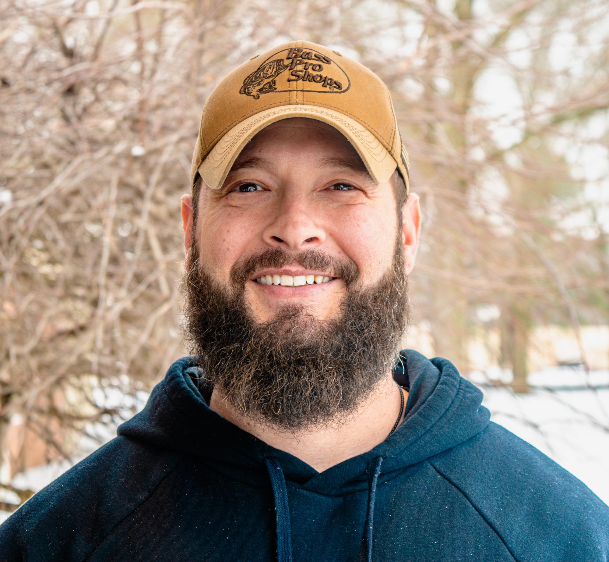 Outdoor portrait of a smiling bearded man in a tan cap and blue hoodie against winter branches