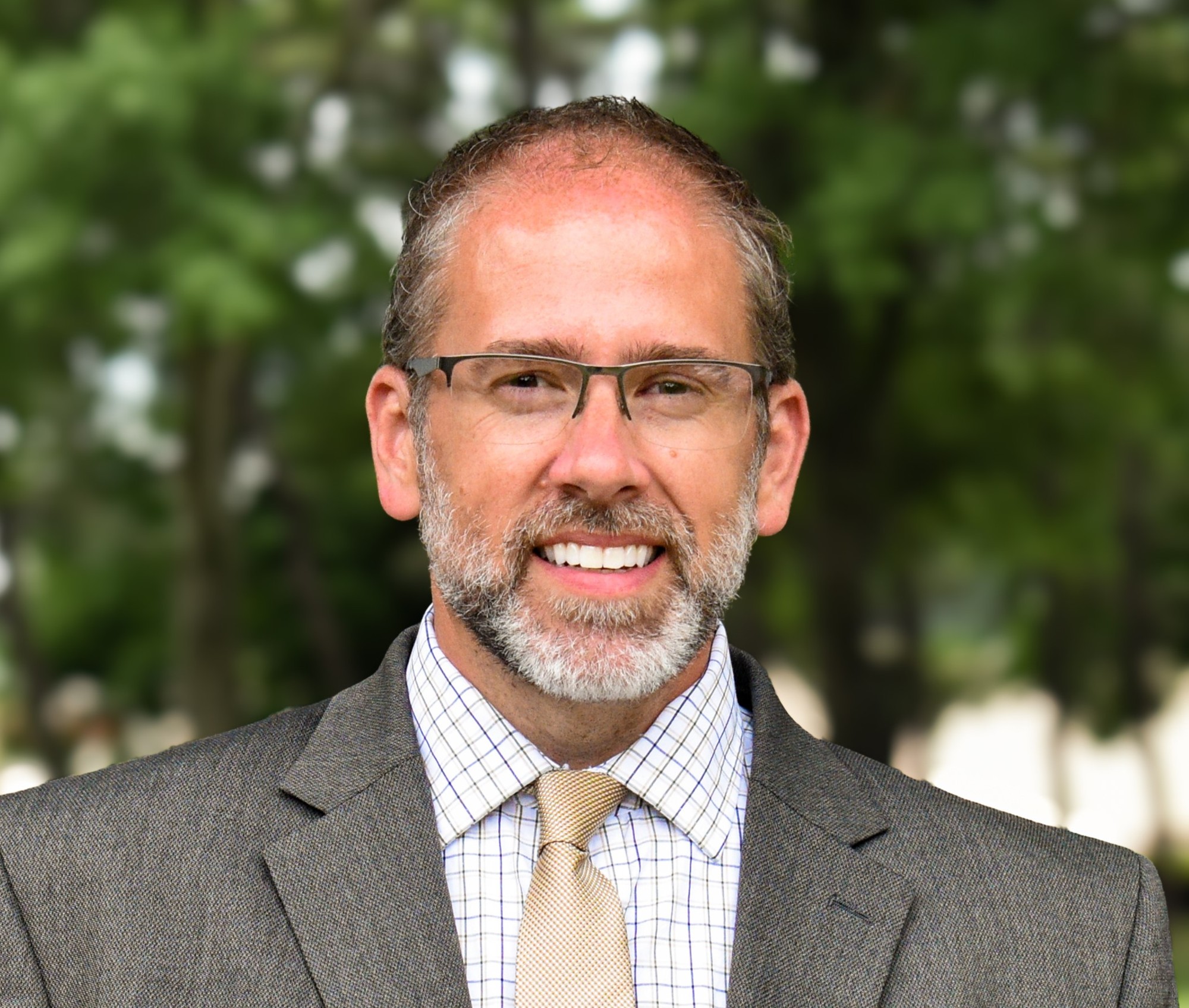 Outdoor professional headshot of a smiling man in glasses, a gray suit, and a gold tie against soft green foliage