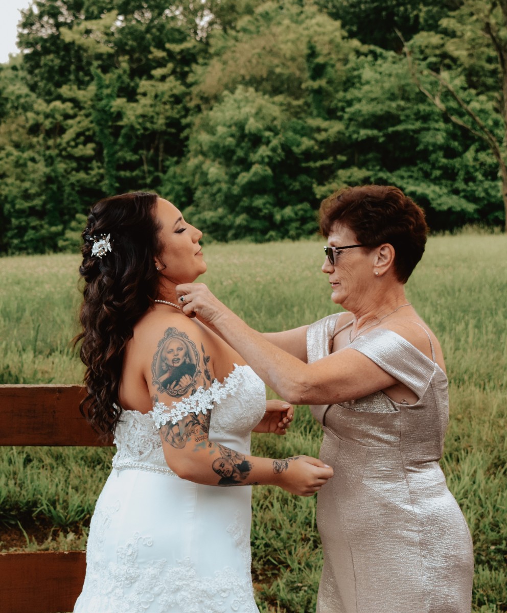 A woman in a silver dress adjusting the bride's necklace beside a wooden fence