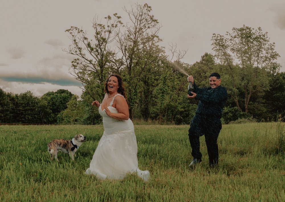 Newlyweds laughing during a champagne spray celebration in a grassy field with their dog
