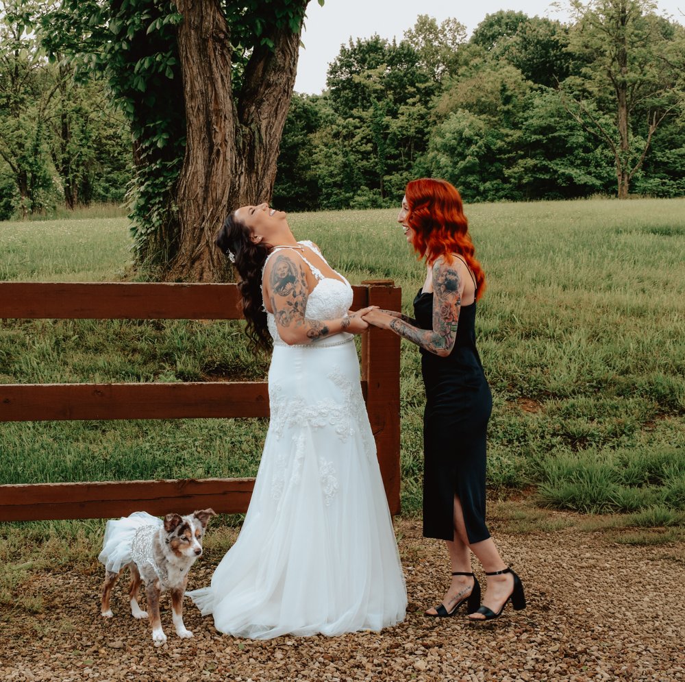 Bride laughing with a red-haired guest beside a wooden fence and their dog