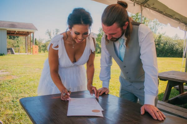 Bride and groom signing their marriage license at a wooden table under the tent