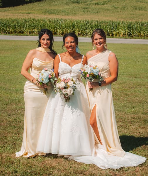 Bride with two bridesmaids in champagne gowns holding pastel bouquets in a green field