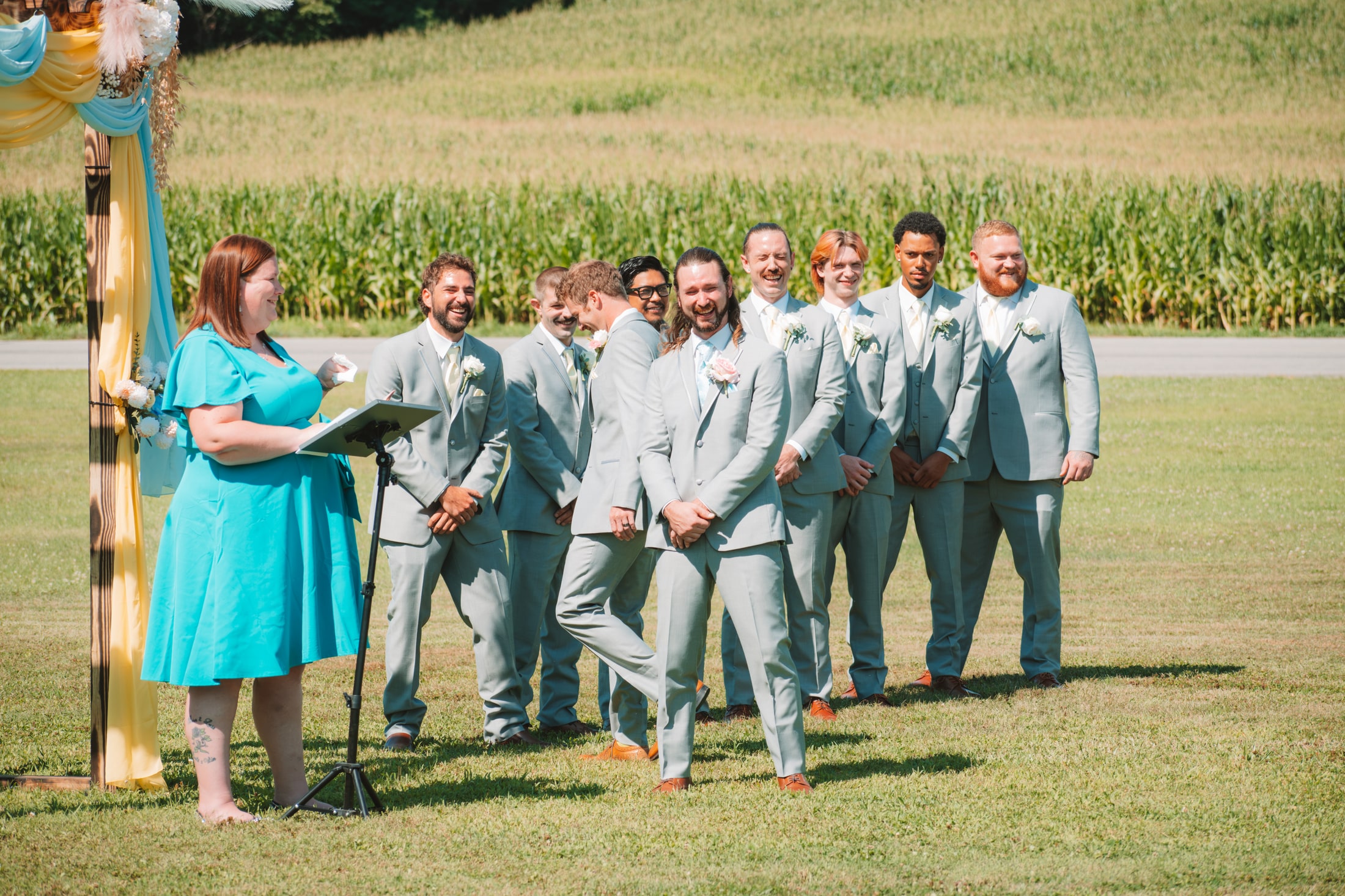 Adult wedding party gathered beside the ceremony arch and cornfield