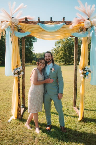 Groom in a sage suit posing with a family member under an arbor draped in yellow and blue fabric